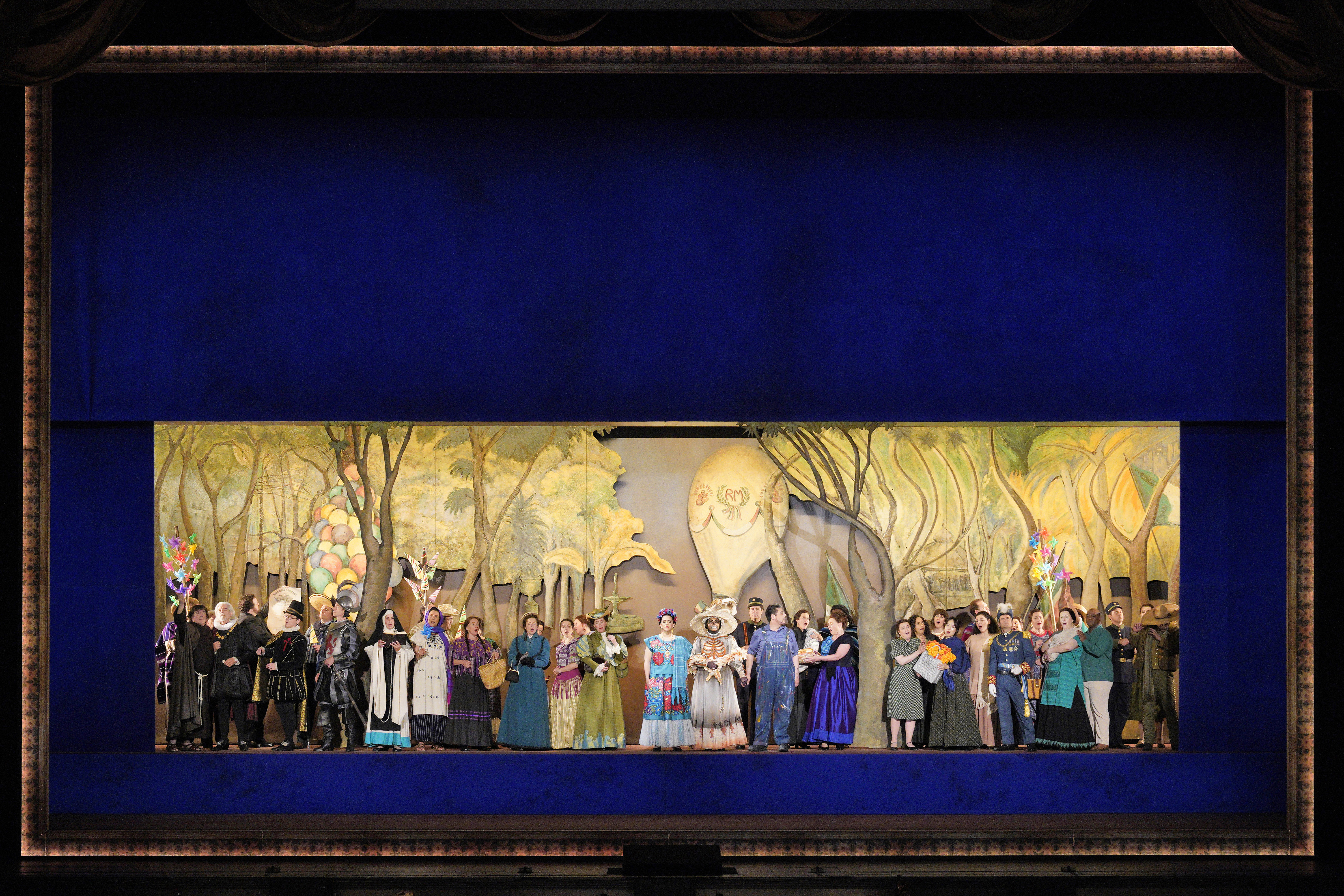 A large ensemble in colorful costumes stands across the stage before a painted forest backdrop, with a skeleton figure near the center.