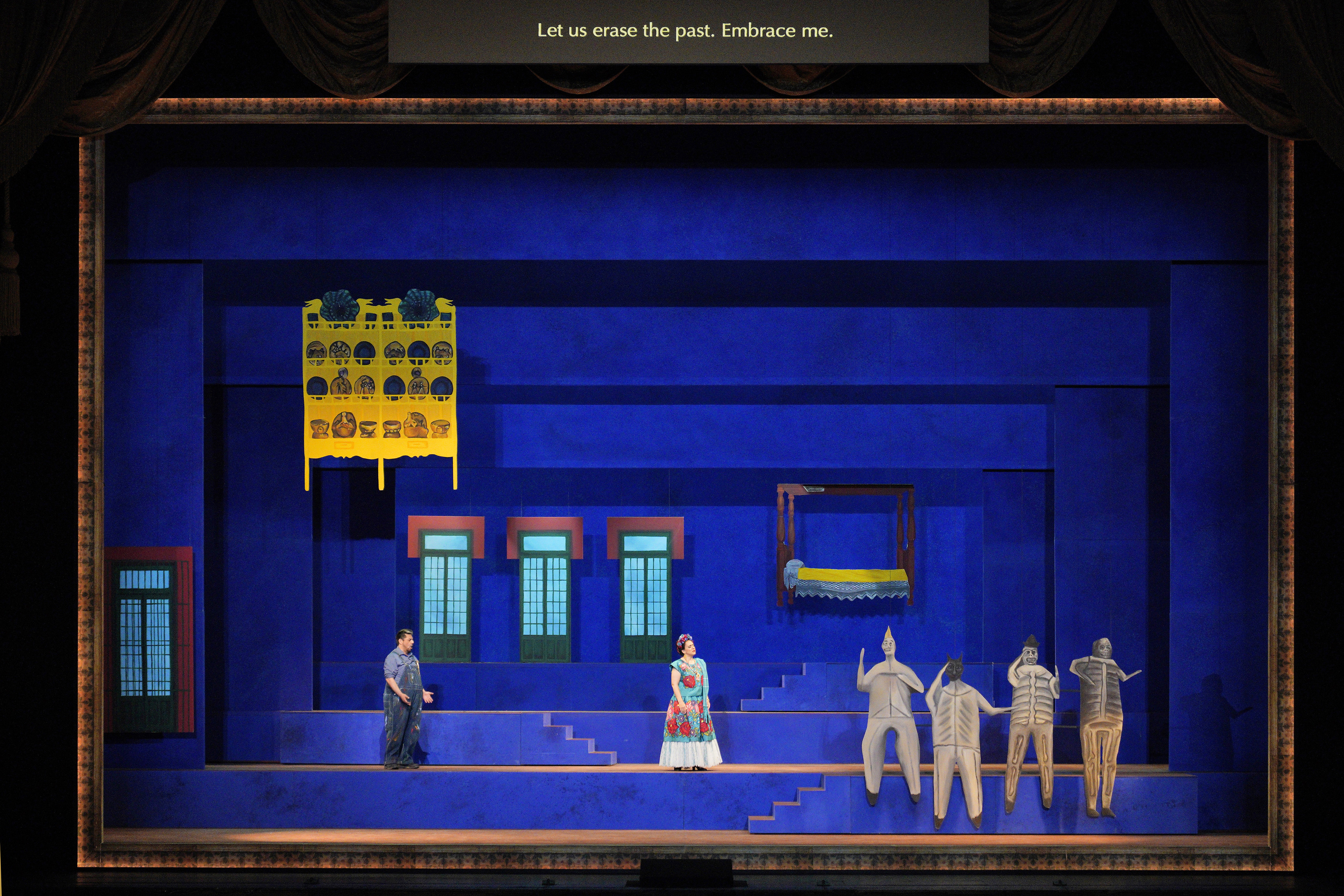A man in paint-splattered overalls faces a woman in a colorful dress inside a blue room, beside four gray sculptural figures.