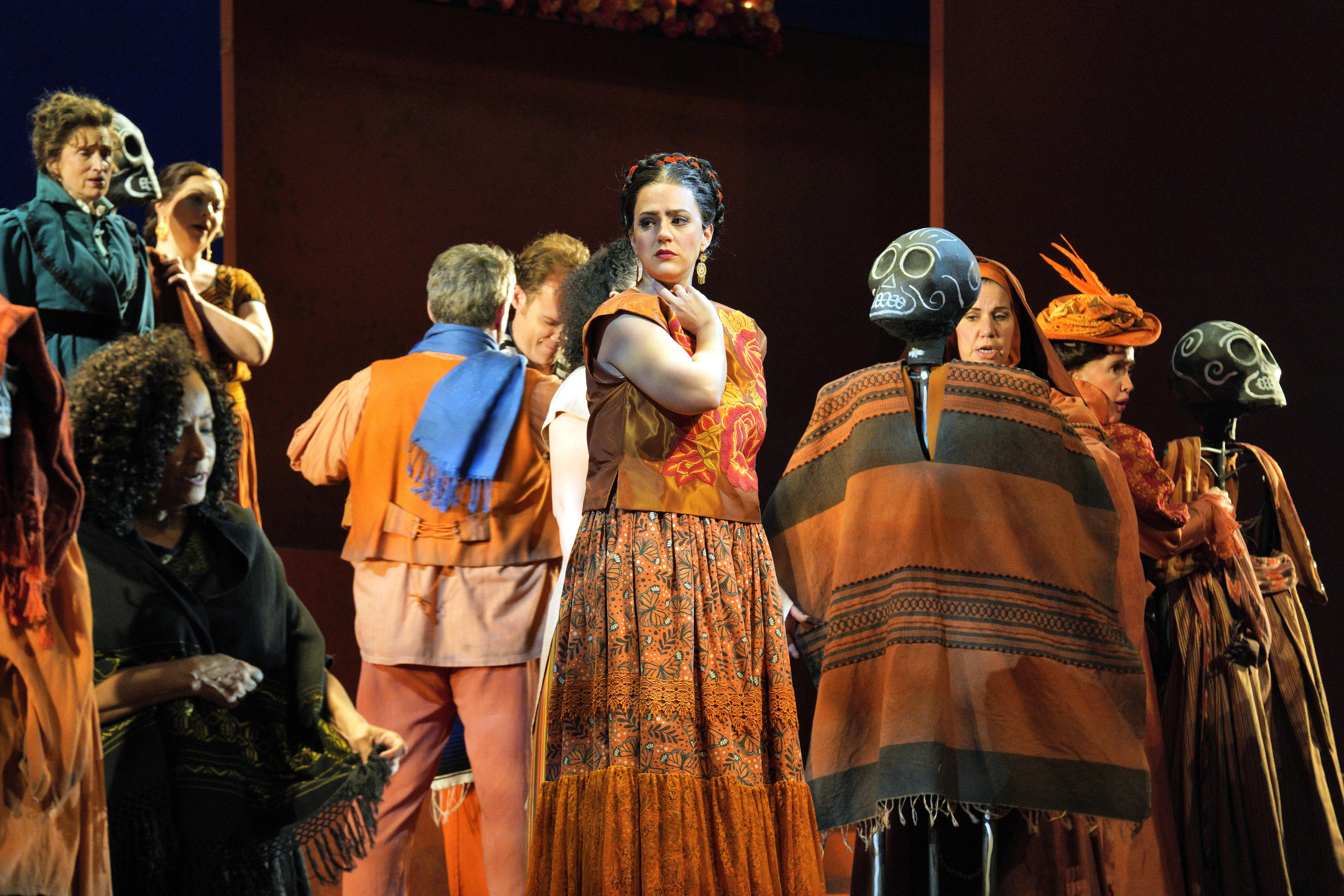 A woman in an orange patterned dress looks over her shoulder while surrounded by performers in colorful costumes and figures carrying black skulls.