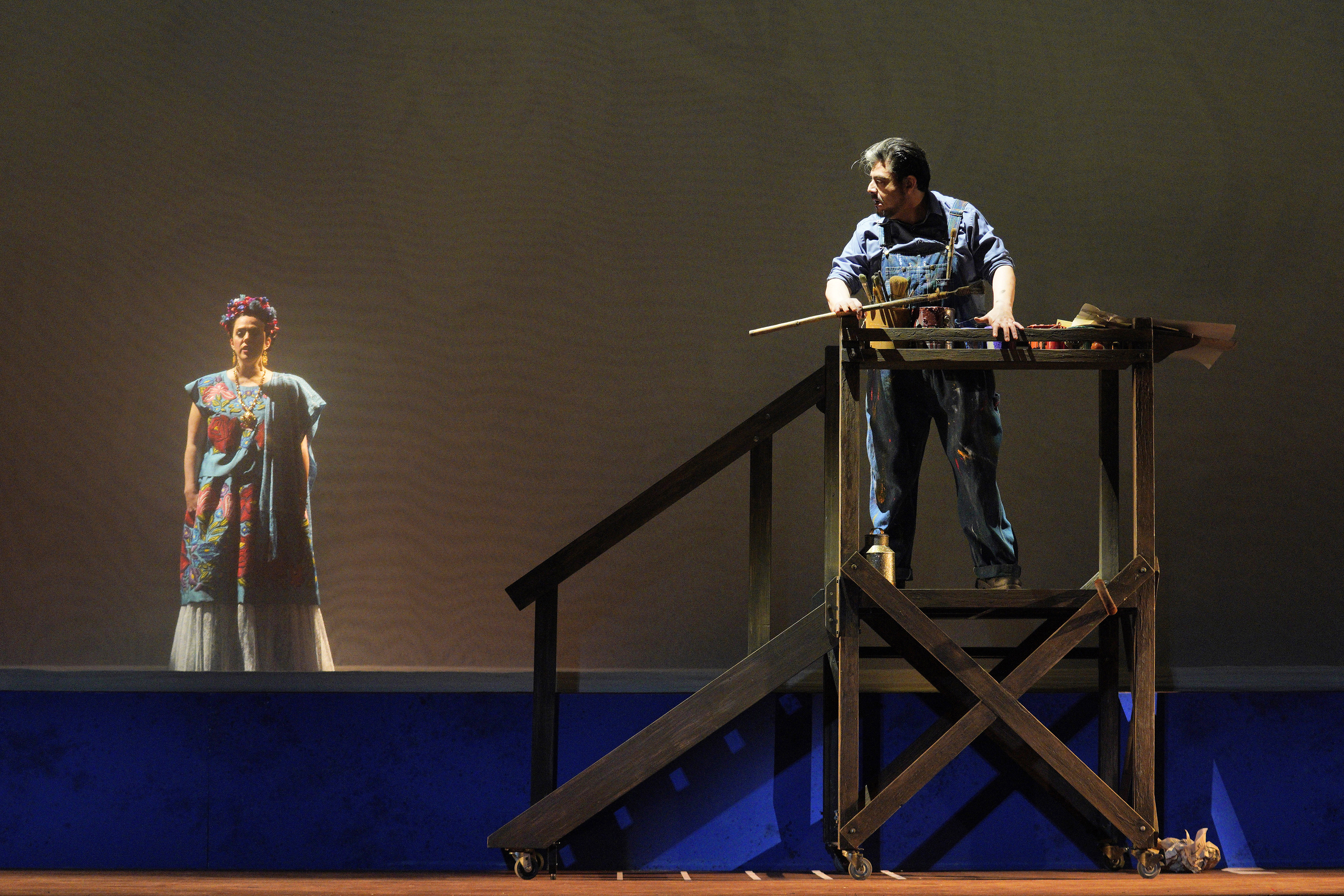 A man in paint-splattered overalls stands at a raised worktable while a woman in a colorful dress watches from below.
