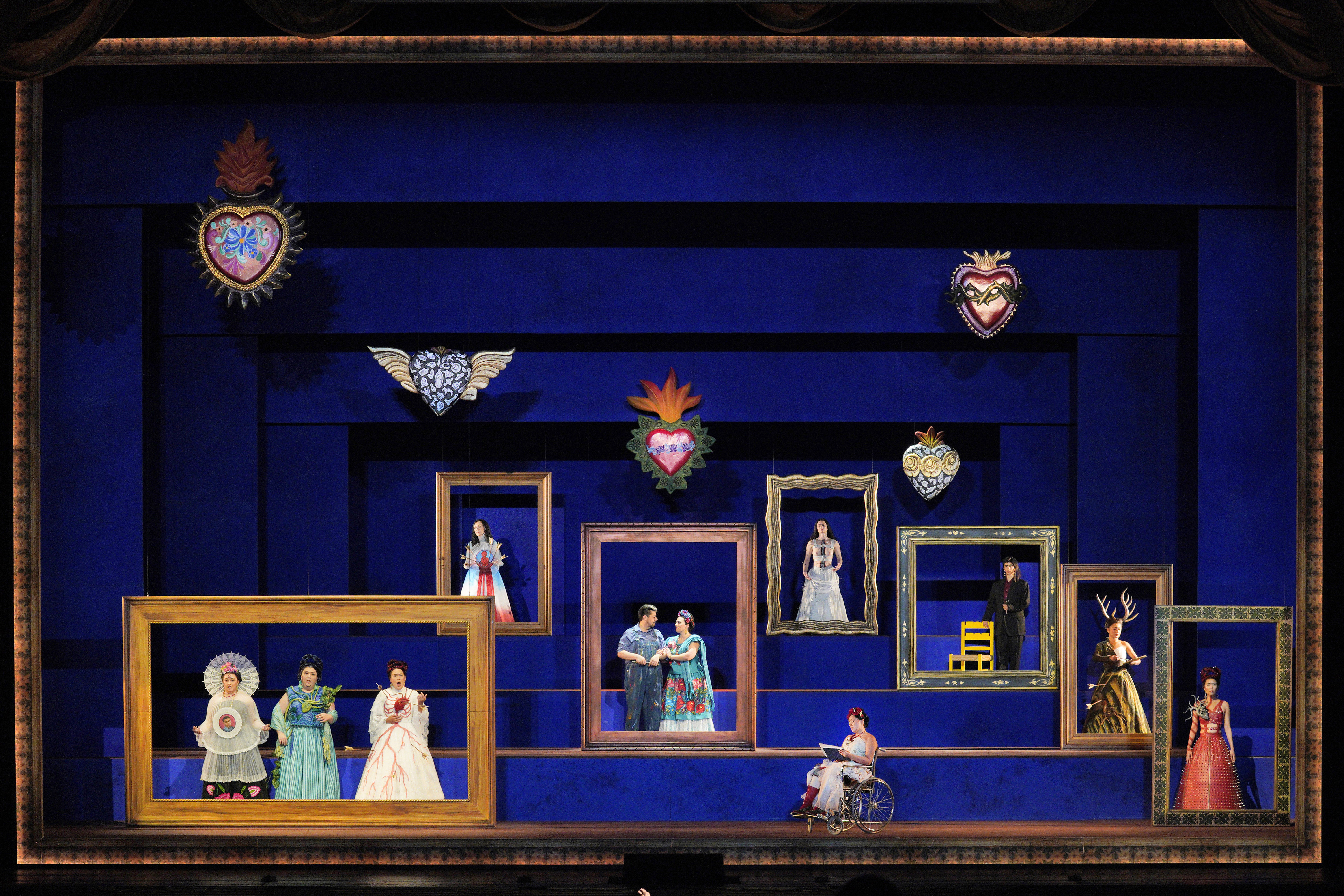Performers in colorful costumes pose within oversized picture frames on a deep blue stage, with a woman seated in a wheelchair below.