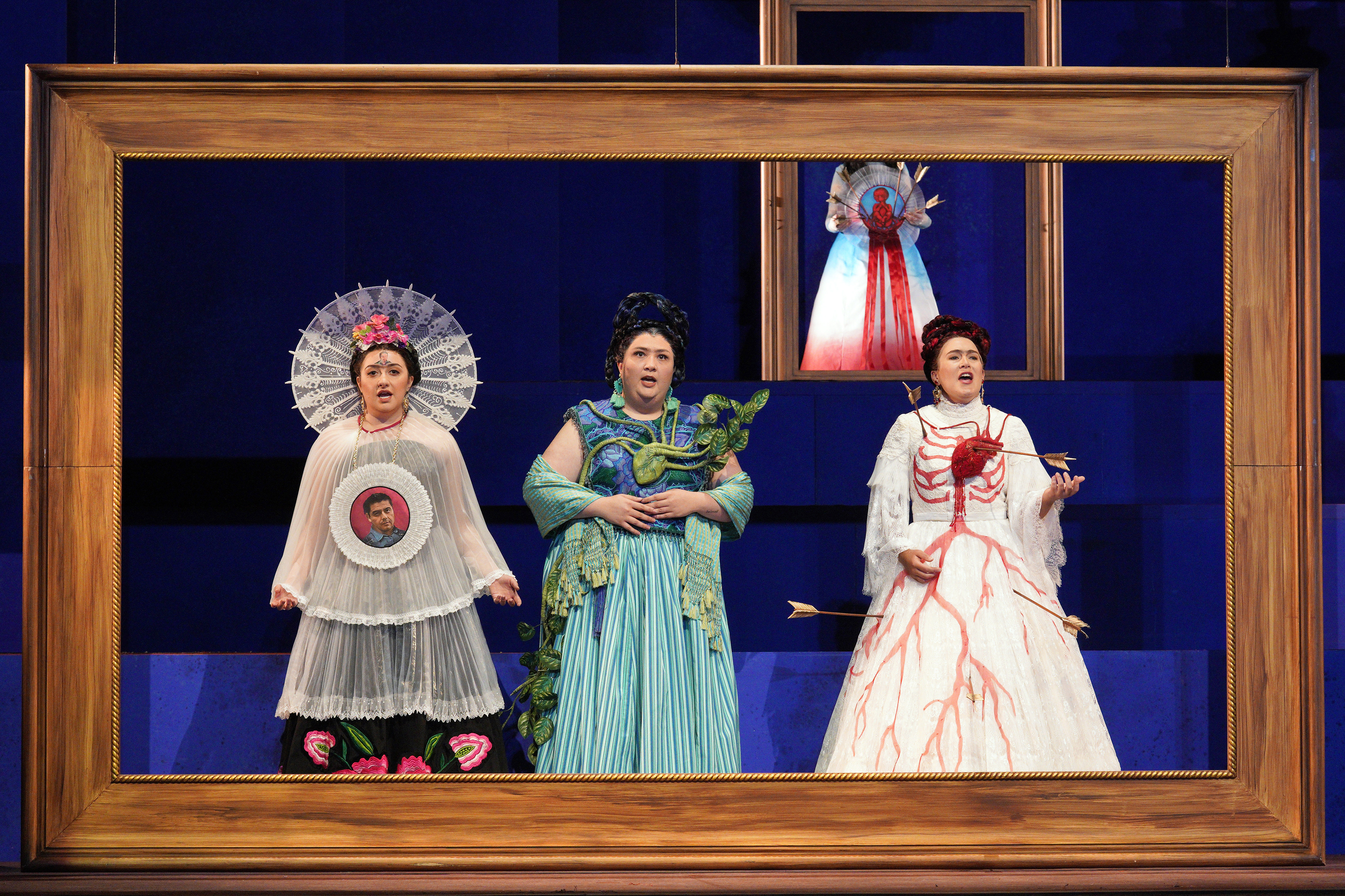 Three women in elaborate symbolic costumes sing within an oversized wooden picture frame, including one in white with a red heart pierced by arrows.