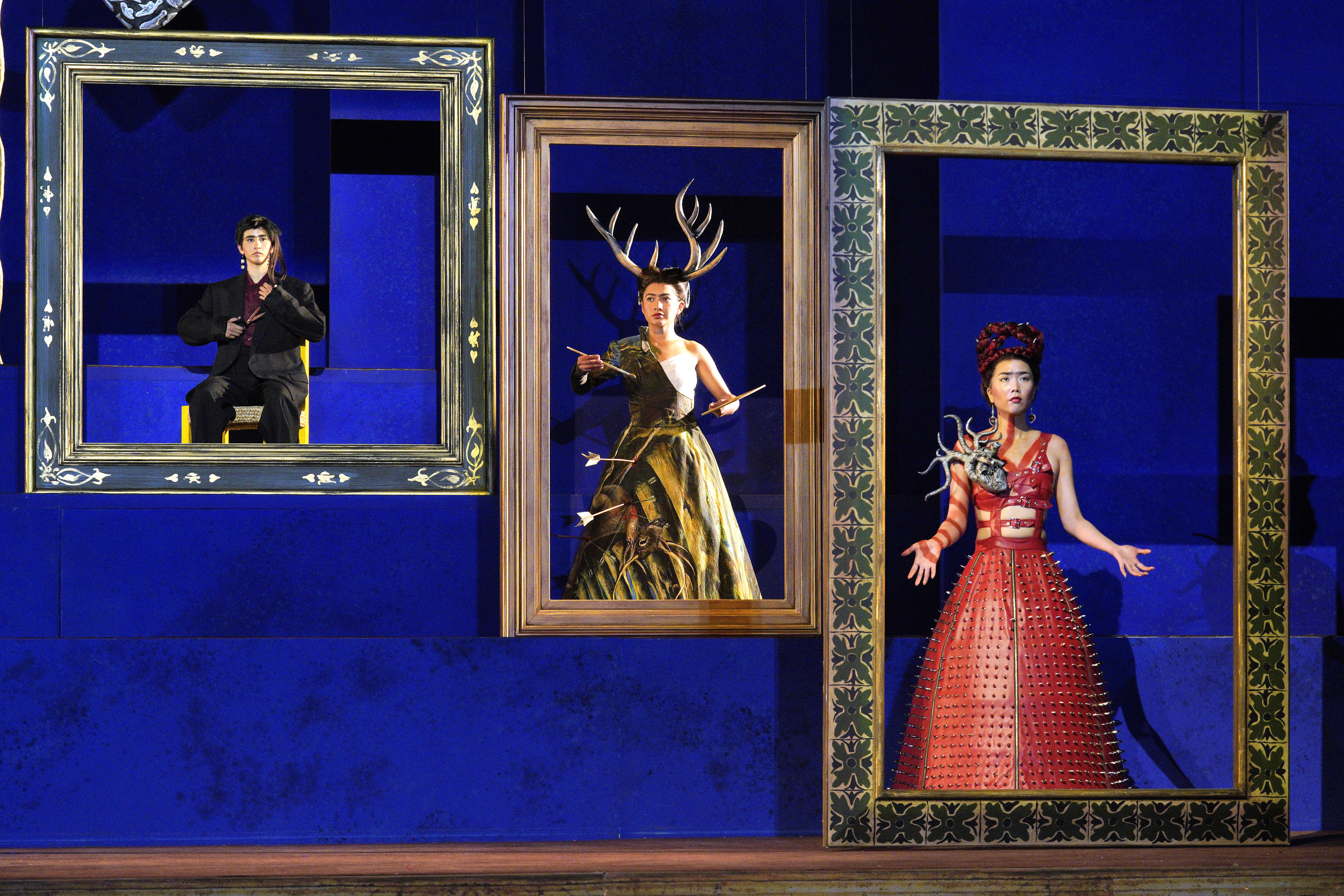Three performers in symbolic costumes stand within oversized picture frames, including one with antlers and arrows and another in a red studded dress.