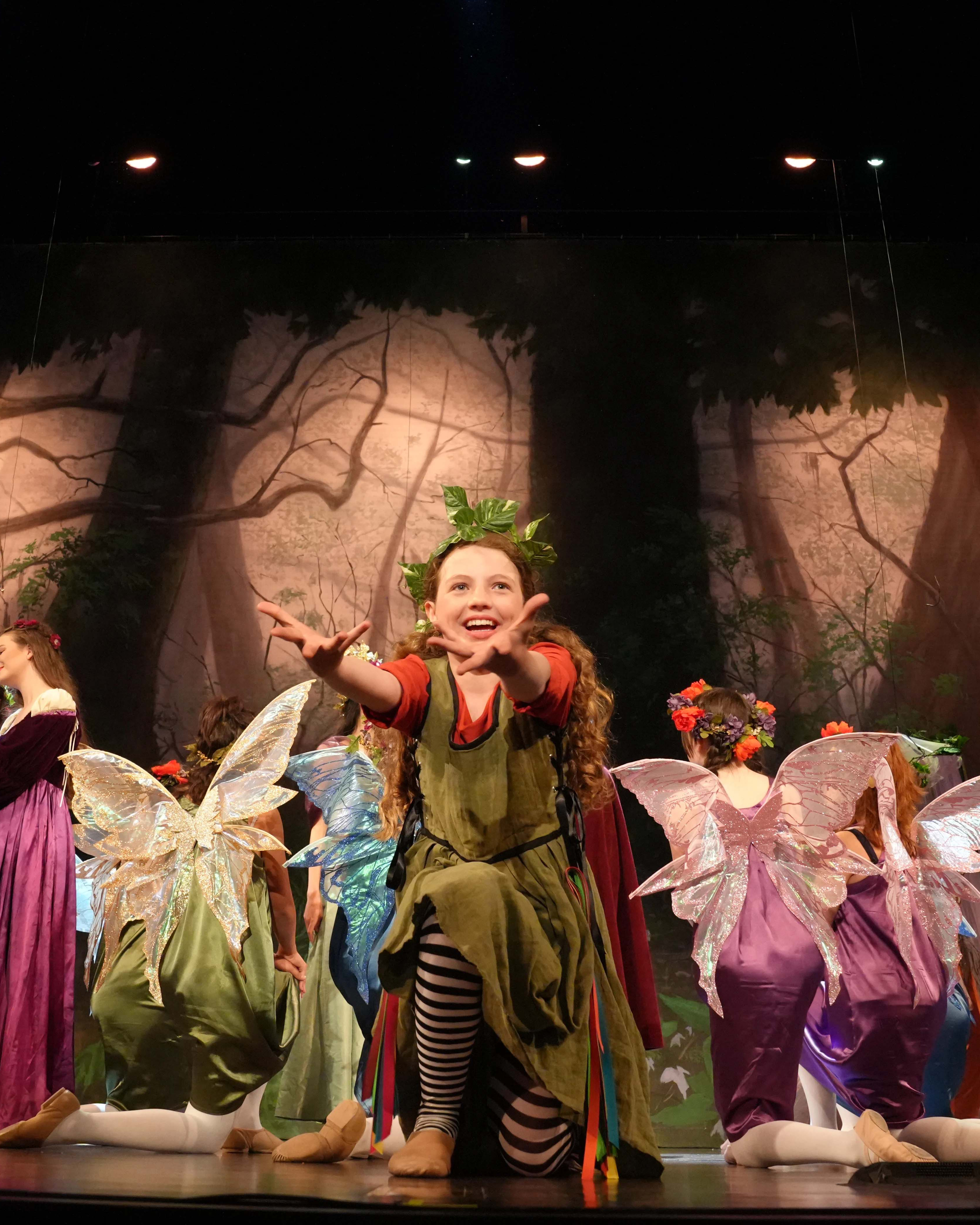 Child performer reaching toward the audience during a forest-themed stage production, surrounded by children dressed as fairies with colorful wings.