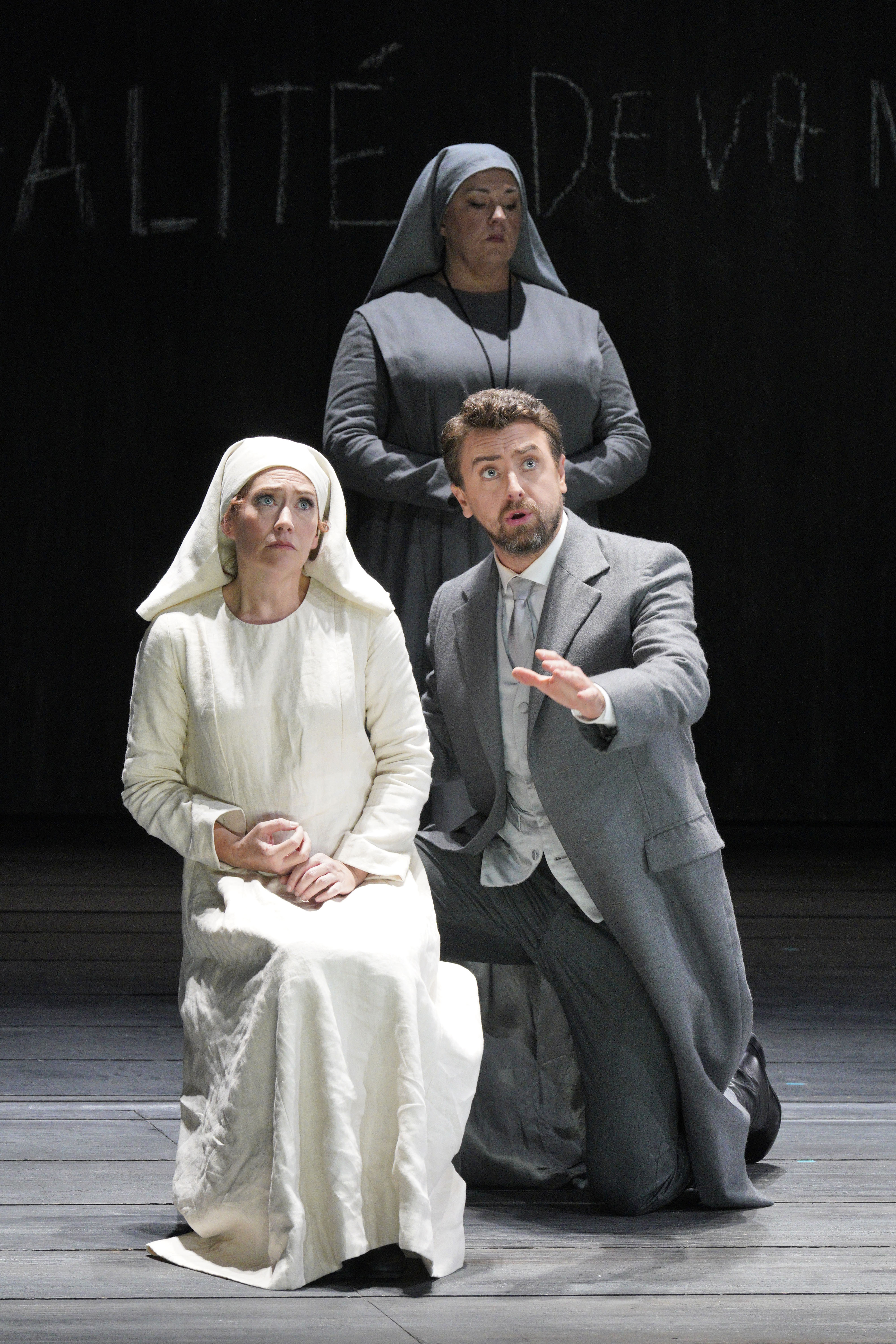 A woman in white sits beside a kneeling man in a gray suit as a nun stands behind them onstage.