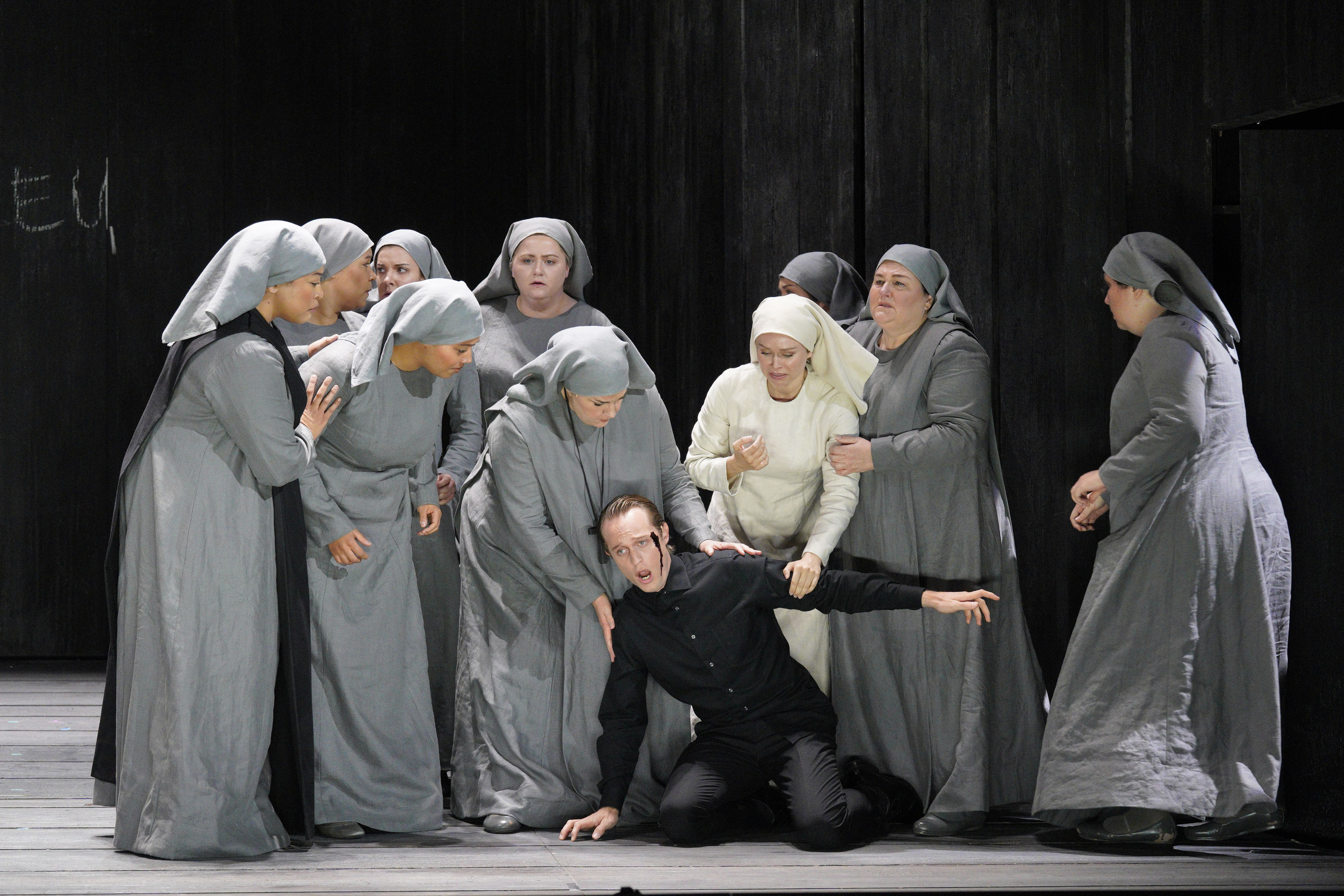 A man in black kneels onstage as nuns in gray habits gather around him and a woman in white supports him.