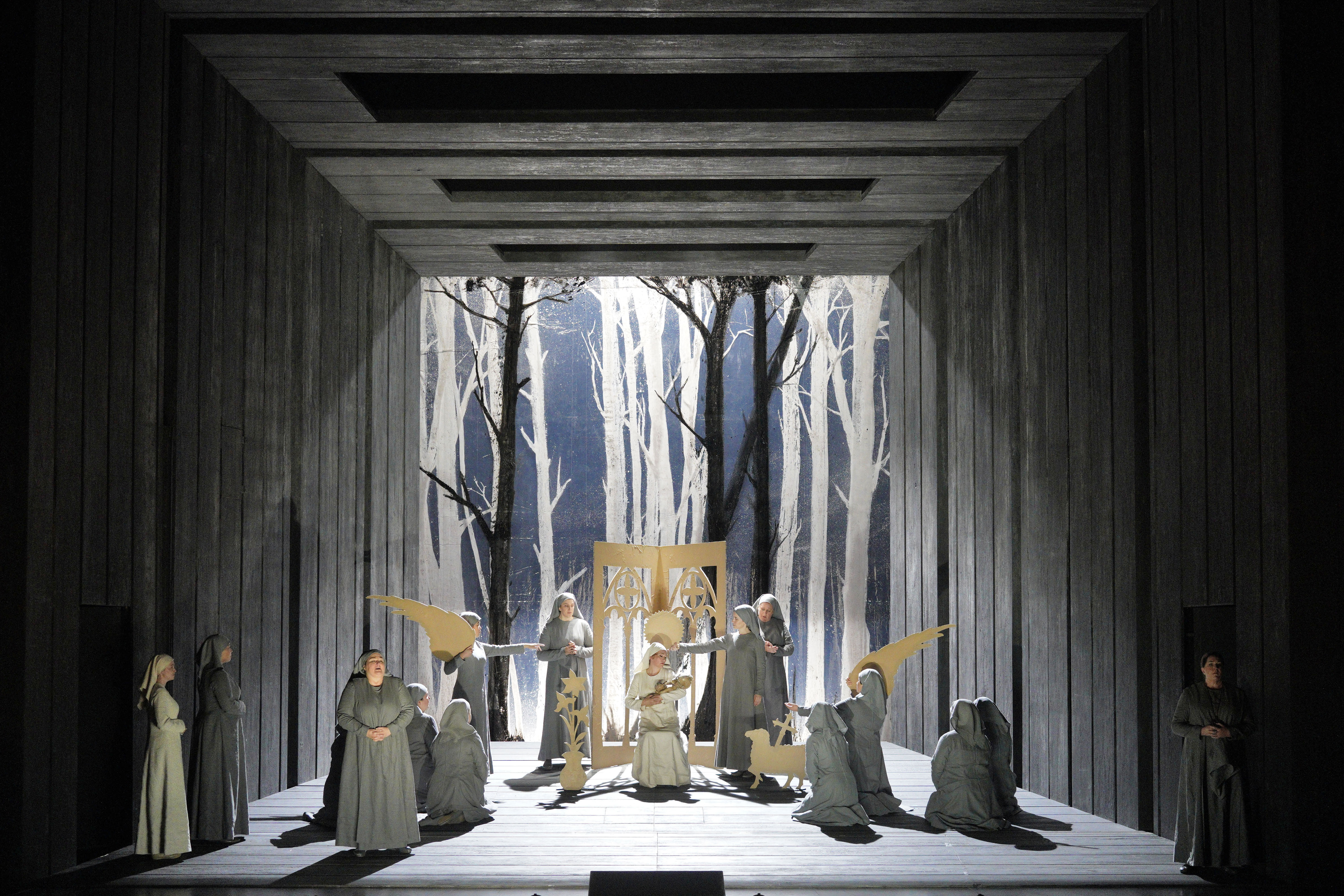 Nuns gather around a seated woman in a forest-like setting framed by a dark wooden corridor.