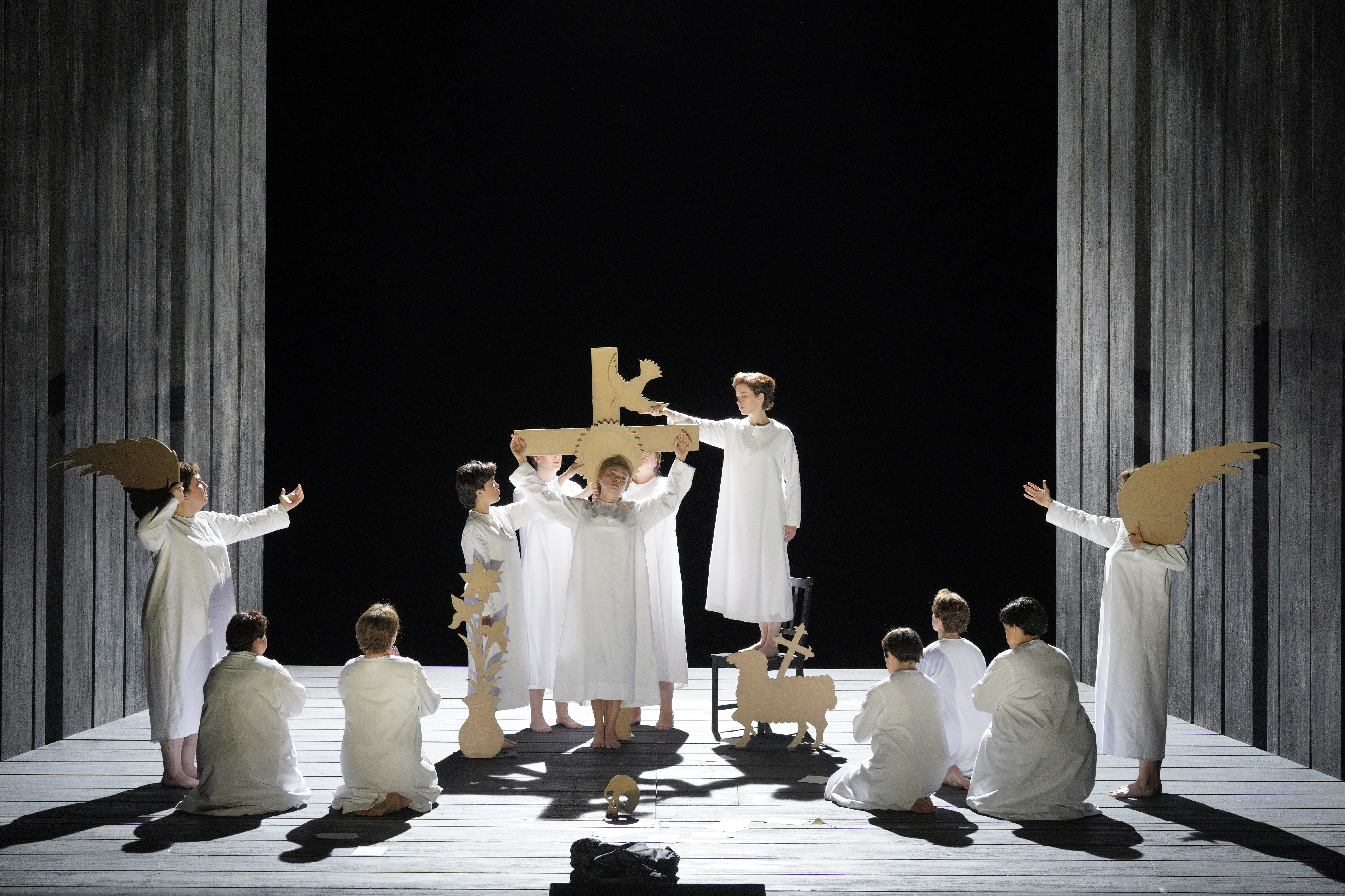 Nuns in white robes gather around a large wooden cross on a stark stage, surrounded by angel, sheep, and dove cutouts.