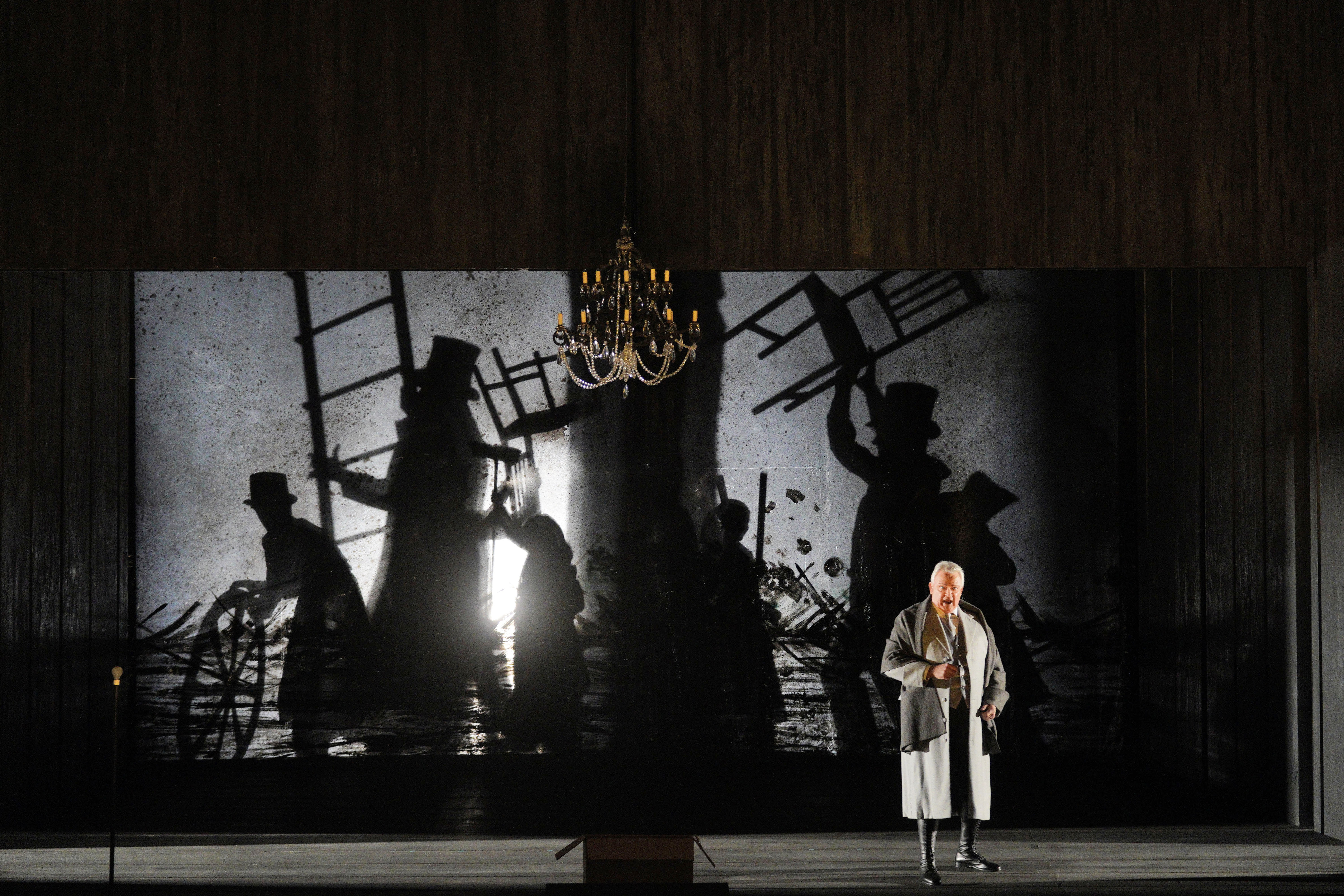 A man in a gray robe stands beneath a chandelier before a projection of silhouetted figures overturning chairs.