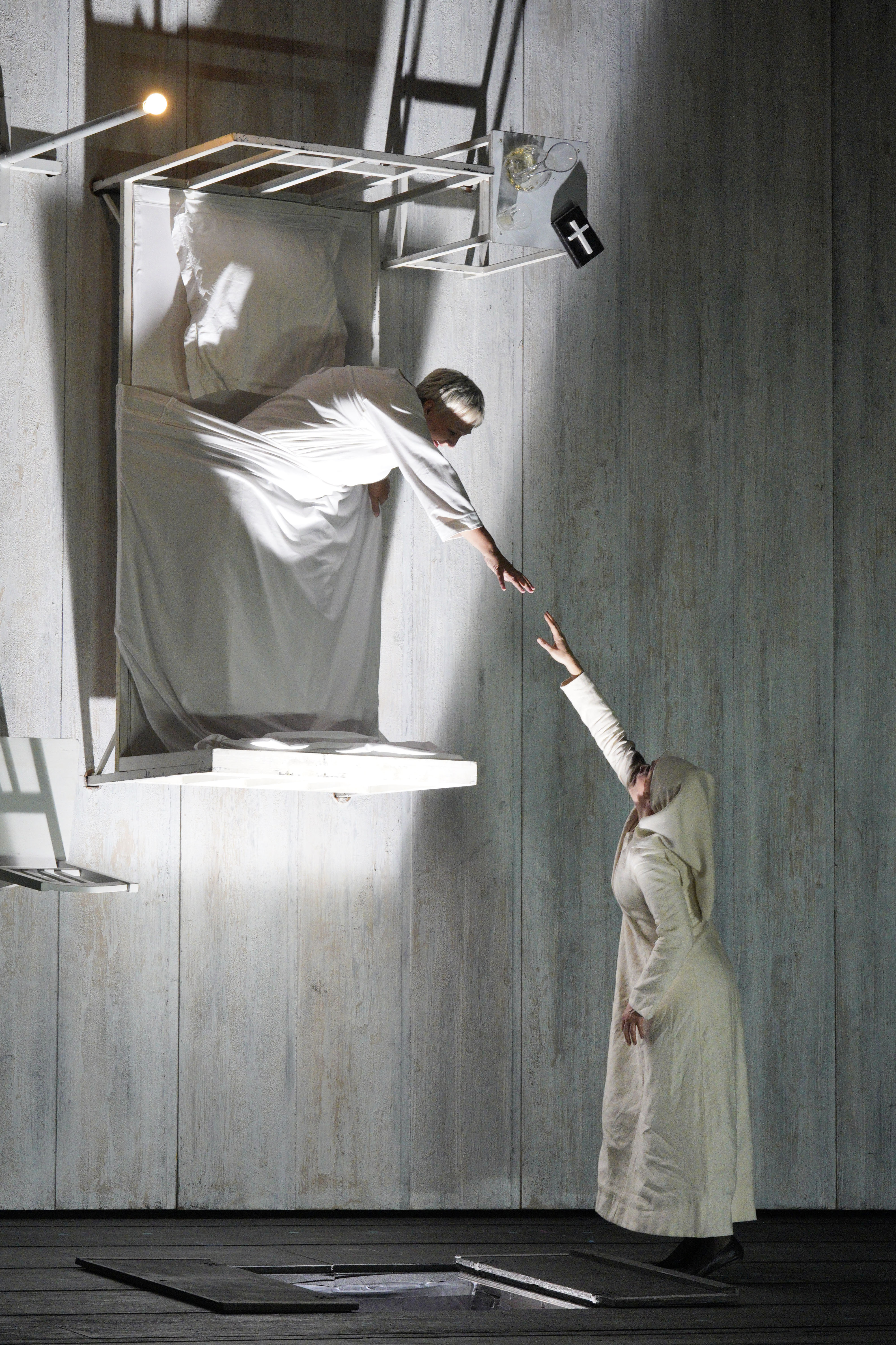 A woman in a suspended bed reaches down toward a nun standing below onstage.